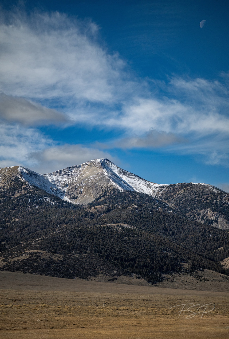 Clouds move over a steep mountain beneath a small moon.