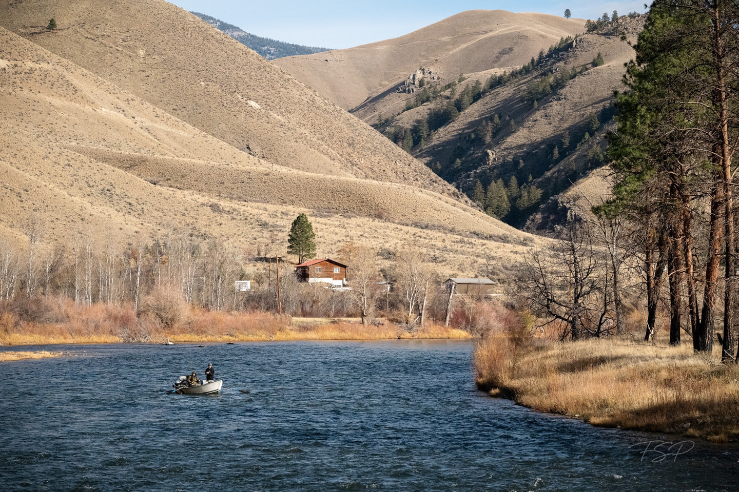 A drift boat travels a Montana river below a cabin and dry hills.